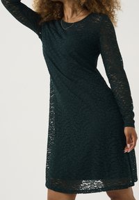 Dark green lace dress with long sleeves, featuring a textured floral pattern and a slightly flared silhouette. Lightweight and delicate fabric.