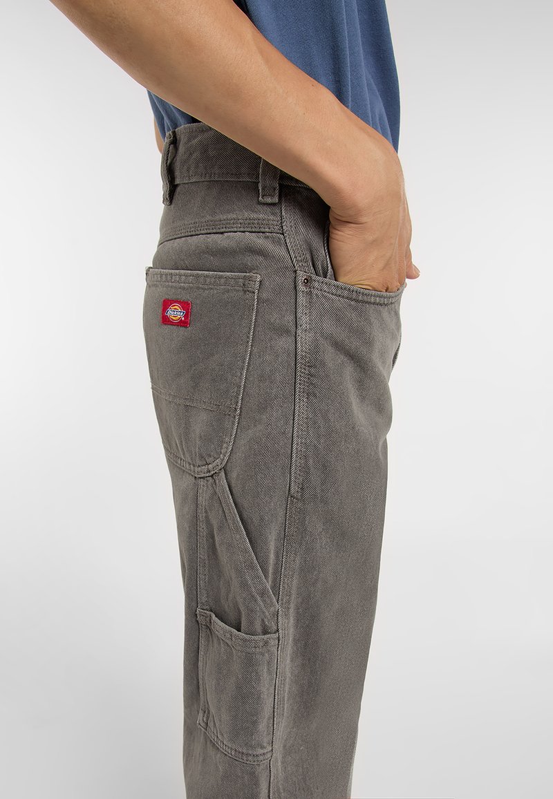 Pantalones de algodón grises con pierna recta, con bolsillos laterales y un parche de logo rojo en el bolsillo trasero. La textura parece resistente.
