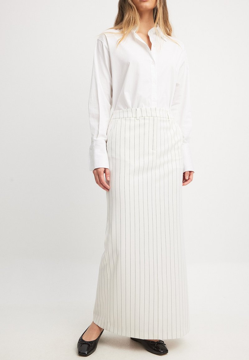 Chemise blanche à manches longues associée à une jupe blanche ajustée taille haute ornée de rayures noires verticales ; des ballerines noires à bouts pointus complètent la tenue.