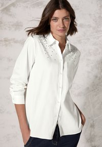 Witte button-up shirt met zilveren accentstuds op de schouders, lange mouwen opgerold bij de manchetten en een relaxte silhouet.