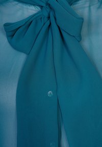 Blusa teal con un grande fiocco nel collo, texture trasparente e piccoli bottoni sul davanti.