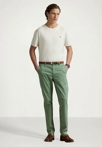 Άνδρας με λευκό κοντομάνικο πουκάμισο, πράσινα παντελόνια και καφέ ζώνη, καφέ επίσημα παπούτσια, στέκεται με τα χέρια στις τσέπες του σε απλό φόντο.