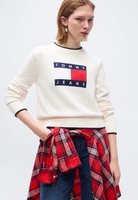 Νεαρή γυναίκα που φοράει άσπρο πουλόβερ Tommy Jeans, κόκκινο καρό πουκάμισο δεμένο στη μέση και σκούρο μπλε τζιν, σε απλό φόντο.