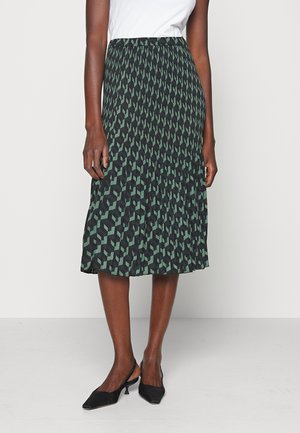 Mujer con falda midi plisada con patrón geométrico negro y verde, camiseta blanca y zapatos slingback negros de tacón bajo, de pie contra una pared blanca.