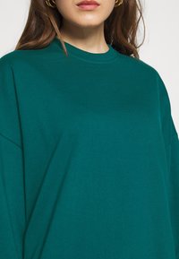 Felpa oversize color aqua con colletto rotondo, realizzata in morbido tessuto di cotone. Caratterizzata da spalle scese e una consistenza liscia. Senza fantasie né dettagli.