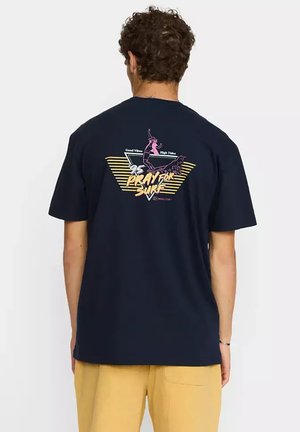 Uomo con capelli ricci che indossa una maglietta blu navy con una grafica colorata di surf e il testo "95 Pray for Surf" sulla schiena, abbinata a pantaloni gialli.