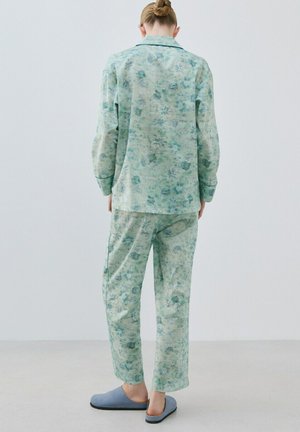 Personne debout, vue de dos, portant un pyjama vert clair à motifs floraux et des chaussons bleu clair, sur un sol neutre avec un fond blanc.