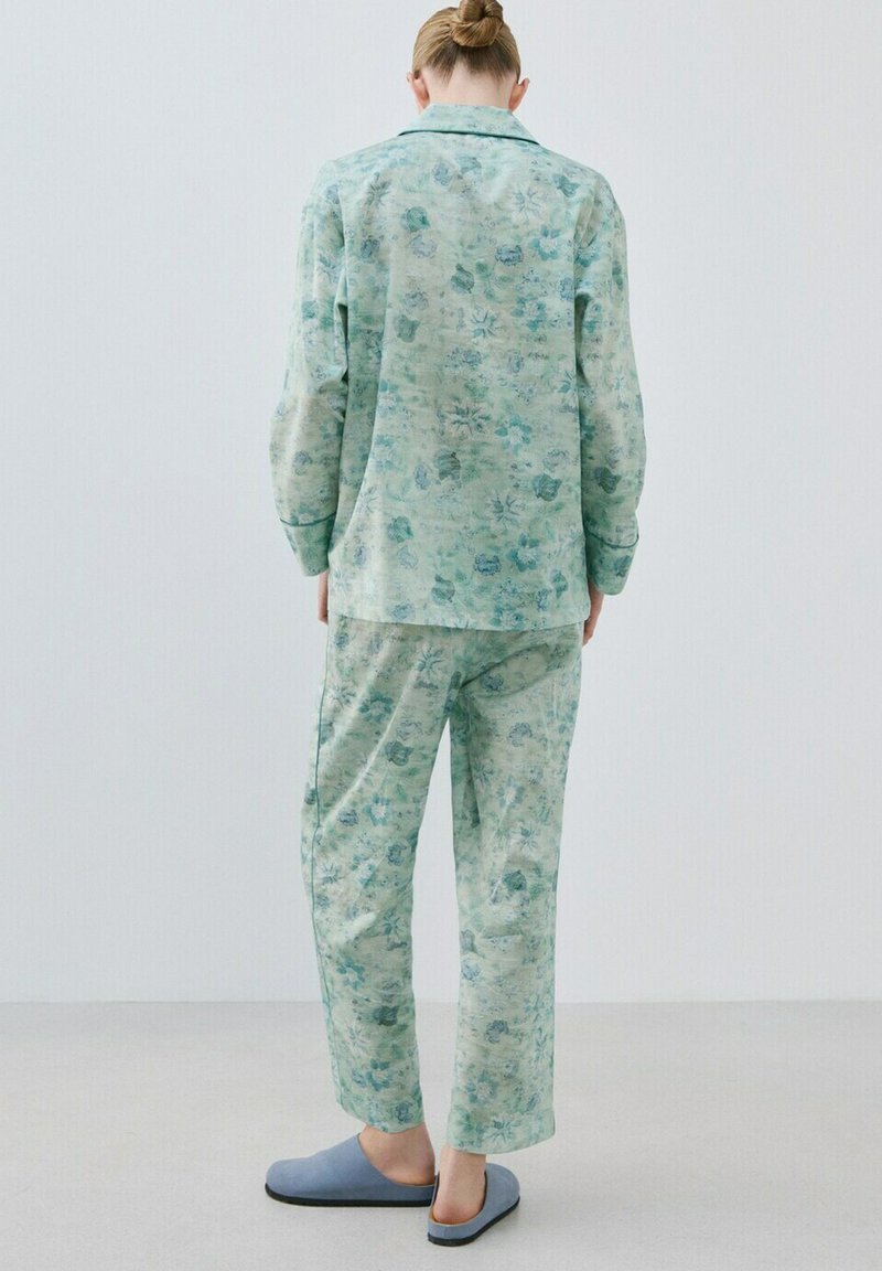 Personne debout, vue de dos, portant un pyjama vert clair à motifs floraux et des chaussons bleu clair, sur un sol neutre avec un fond blanc.