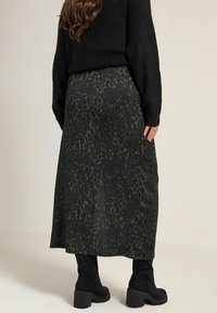 Musta neulottu neule pitemmän tummanvihreän hameen päällä, jossa on hienovarainen leopardikuvio. Mallilla on jalassa polvimittaiset mustat saappaat.