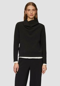 Schwarz langärmliges Oberteil mit Cowl-Ausschnitt. Der Stoff wirkt glatt und weich. Darüber getragen ist ein weißes Hemd mit ausgefranstem Saum.