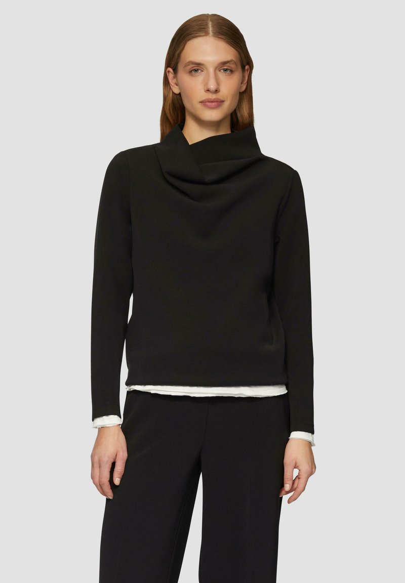 Schwarz langärmliges Oberteil mit Cowl-Ausschnitt. Der Stoff wirkt glatt und weich. Darüber getragen ist ein weißes Hemd mit ausgefranstem Saum.