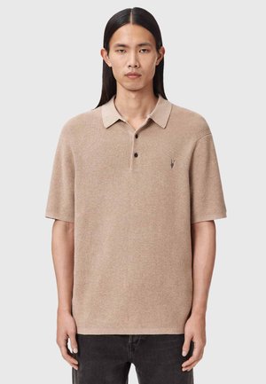 Mand med langt sort hår iført en beige kortærmet strikket polotrøje og mørke bukser, der står foran en lysegrå baggrund.