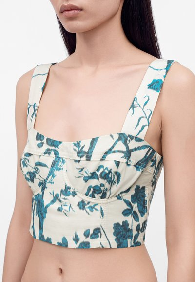 Γυναίκα που φοράει λευκό crop top με φαρδιές τιράντες και μπλε floral σχέδιο, που δείχνει τους ώμους και το άνω μέρος του κορμού.