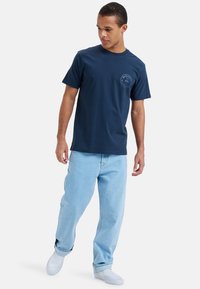 Camiseta de algodón azul marino de manga corta, con un diseño de logo circular en el lado izquierdo del pecho. Llevada con jeans de color azul claro y zapatillas blancas.