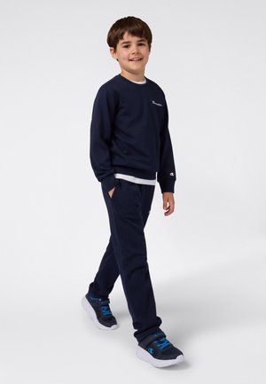 Marineblauwe trui en bijpassende broek van zachte stof, met een logo op de borst en elastische manchetten. Gecombineerd met sportieve grijze schoenen.