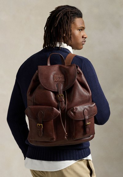 Man bär marinblå tröja, vit skjorta och beige byxor, bär stor brun läderryggsäck med fickor framtill med spänne.