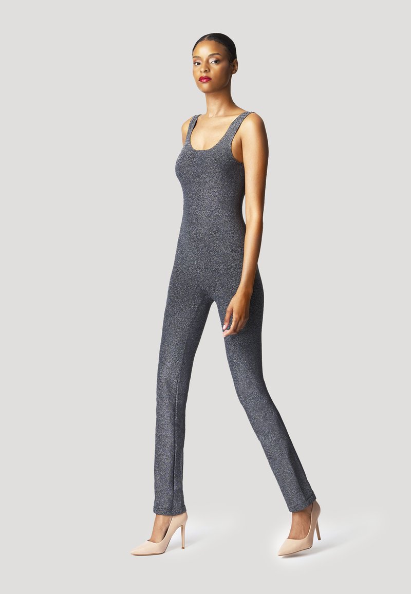 Jumpsuit gris scintillant au design ajusté, avec de larges bretelles et des jambes évasées, associé à des chaussures à talons nus.