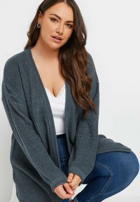 Cardigan grigio lavorato a maglia con apertura frontale, maniche lunghe e polsini a coste. Indossato su una maglietta bianca, con un motivo texturizzato e una vestibilità morbida.