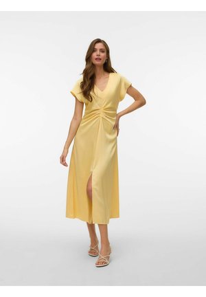 Žena v žltej midi šatách s lietavými rukávmi, záhybmi vpredu a predným rozparkom, nosi béžové sandále na opätku na bielom pozadí.