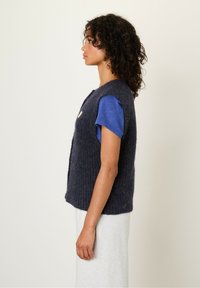 Vrouw met krullend haar, gekleed in een marineblauwe mouwloze wollen vest over een blauwe shirt met korte mouwen en een lichtgrijze rok, staande in profiel.