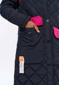 Ruka osoby sklouzává jasně růžovou peněženku do velké černé prošívané kapsy na bundě s knoflíkem a přišitými štítky.