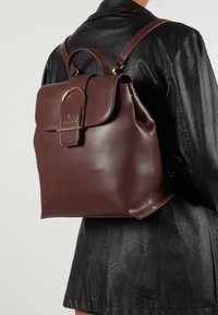 Brun läderryggsäck med en strukturerad form, med en vikbar klaff och en guldfärgad hårdvarudetalj. Slät, elegant textur.