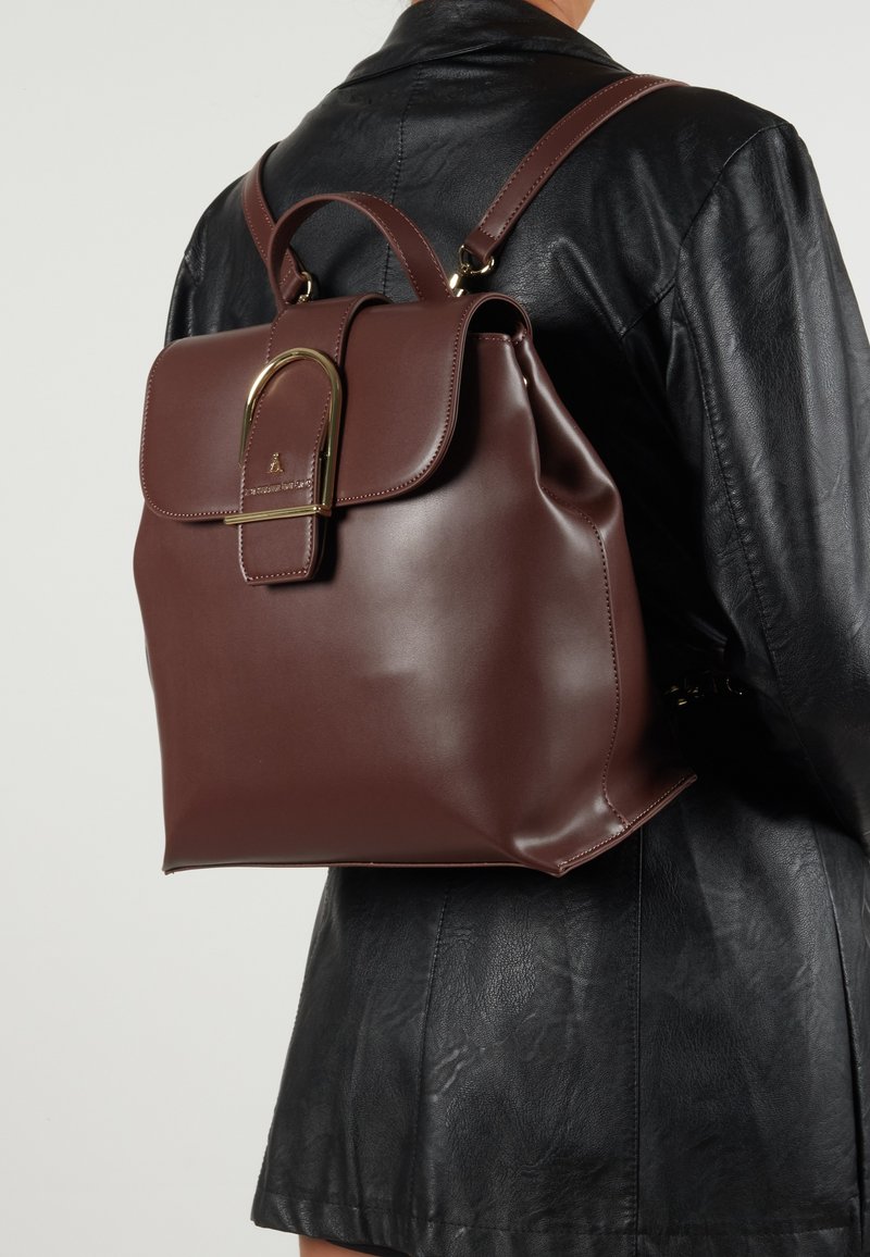 Brun läderryggsäck med en strukturerad form, med en vikbar klaff och en guldfärgad hårdvarudetalj. Slät, elegant textur.