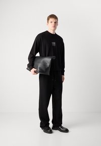 Zwart sweatshirt en broekset, met een getextureerde patch op de borst. Het model houdt een zwarte leren tas met ronde hoeken vast.