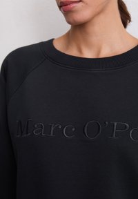 Femme portant un sweat-shirt noir avec un col côtelé et un logo "Marc O'Polo" ton sur ton en relief sur la poitrine.