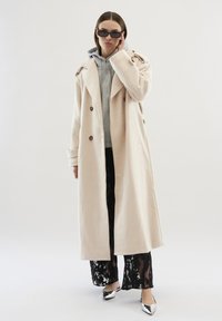 Palton bej de tip trench cu design cu două rânduri de nasturi, având nasturi maro, purtat peste un hanorac gri și pantaloni negri din dantelă, asortați cu pantofi argintii.