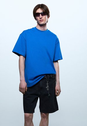 Mladý muž s čiernymi slnečnými okuliarmi, modrým oversize tričkom a čiernymi denimovými kraťasmi s príveskom na kľúče stojaci pred jednoduchým svetlým pozadím.