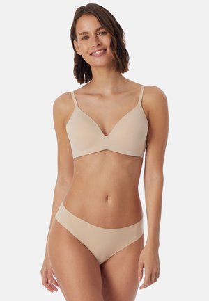 Frau lächelnd, trägt einen beigen nahtlosen BH und passende hochgeschnittene Unterhose, steht vor einem schlichten hellen Hintergrund.