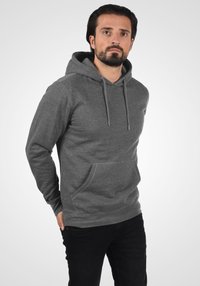 Haut à capuche gris en matériau doux, doté d'une poche avant, d'une capuche avec cordons de serrage et de poignets côtelés. Assorti à un jean noir.