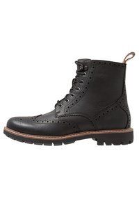 Stivale alla caviglia in pelle nera con dettagli brogue, chiusura con lacci nella parte anteriore e suola in gomma con battistrada testurizzato. Cuciture marroni accentuano il bordo.