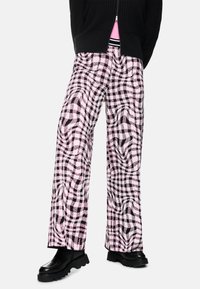 Person, der schwarze klobige Schuhe, weit geschnittene Hosen mit einem pink-schwarzen verzerrten Karomuster und einen teilweise zugeknöpften schwarzen Pullover über einem pinken Oberteil trägt.