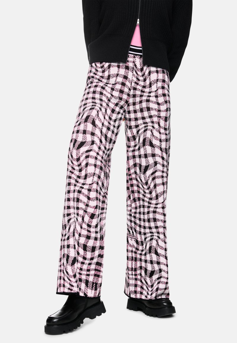 Person, der schwarze klobige Schuhe, weit geschnittene Hosen mit einem pink-schwarzen verzerrten Karomuster und einen teilweise zugeknöpften schwarzen Pullover über einem pinken Oberteil trägt.