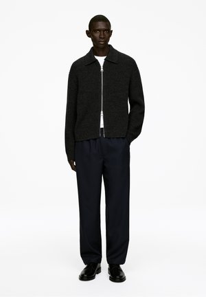 Schwarze, gerippte Zip-Jacke mit Kragen, kombiniert mit marineblauen Hosen und schwarzen Schuhen. Einfaches Design, strukturiertes Material und neutrale Farben.