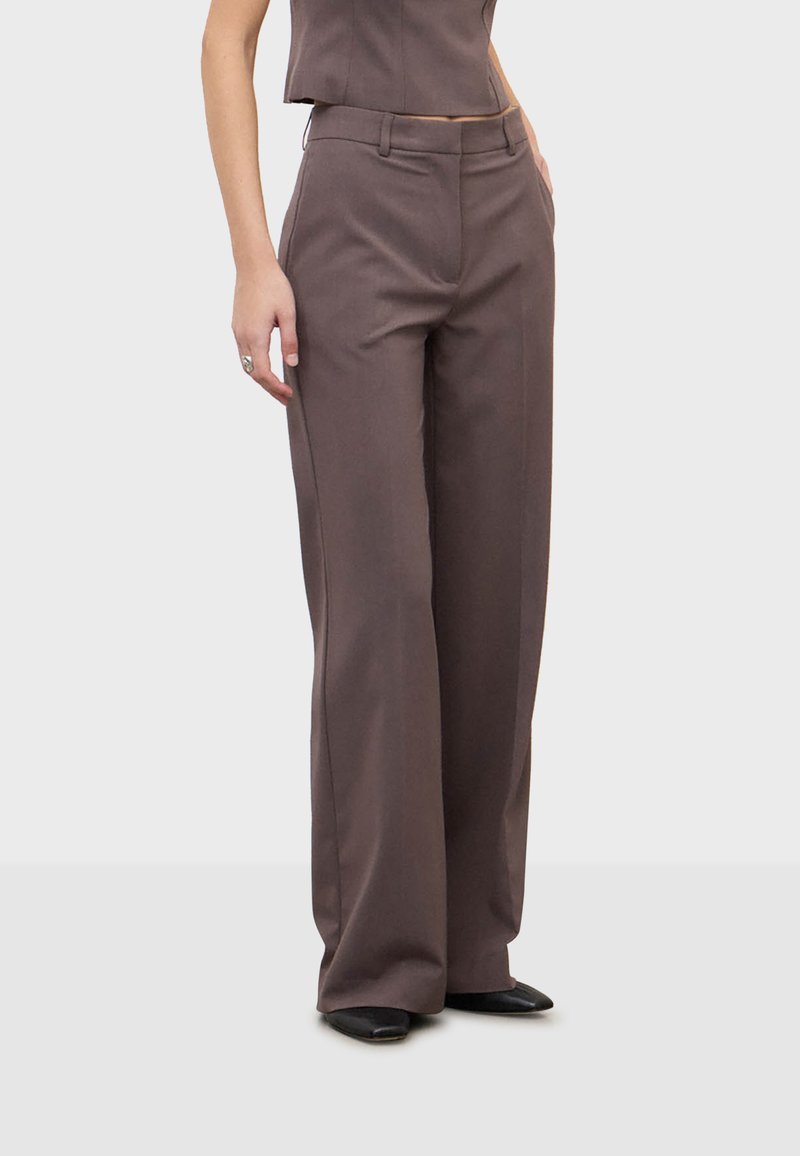 Femme portant un pantalon marron taille haute à jambes larges avec des poches latérales et des chaussures noires à bout pointu, debout devant un fond uni.