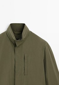 Giacca verde oliva con colletto alto, chiusura frontale a scatto, due tasche laterali e una texture del tessuto liscia e leggera.