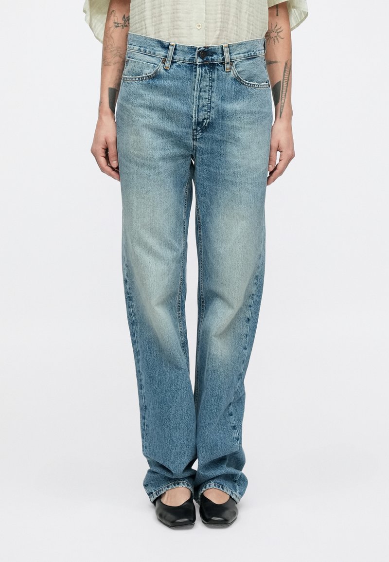 Világoskék, egyenes szárú farmer, fakó farmeranyagból; öt zsebbel, gombos záródással és varrásdíszítéssel. Fekete cipővel viselve.