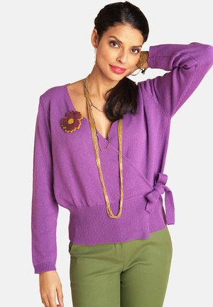 Frau trägt einen lila Wickelpullover mit einer Blütenbrosche und einer goldenen, mehrlagigen Halskette, kombiniert mit olivgrünen Hosen, posiert mit der Hand hinter dem Kopf.