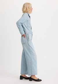 Jasnoniebieski denimowy kombinezon z długimi rękawami, przednimi kieszeniami i szerokimi nogawkami, połączony z czarnymi płaskimi butami z małym akcentem.