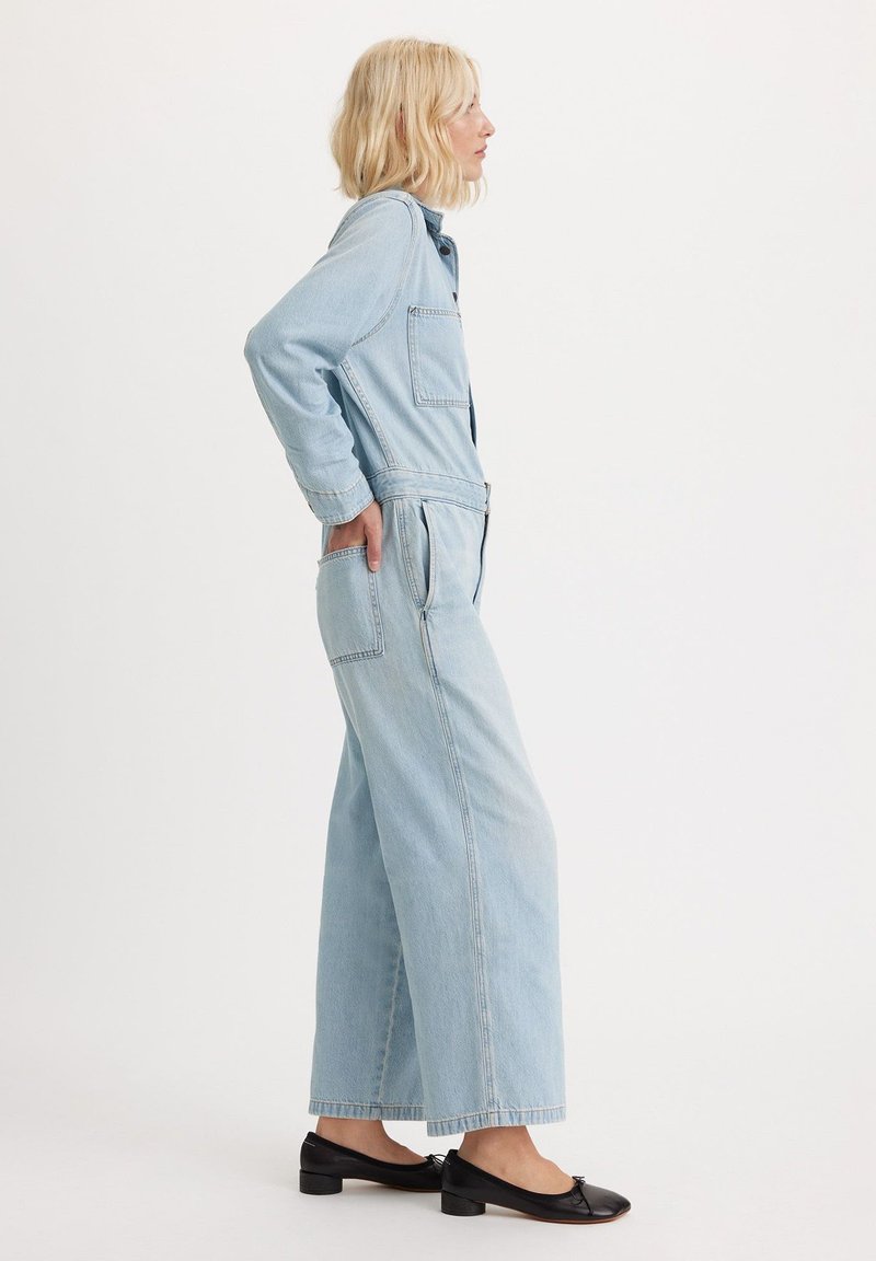 Lyseblå denim-dragt med lange ærmer, forlommer og vide ben, kombineret med sorte flade sko med en lille detalje.