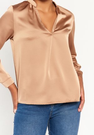 Kvinde iført en skinnende beige satin V-hals bluse med lange ærmer kombineret med blå denimjeans.