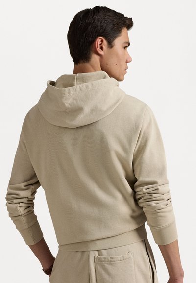Beige huppari, jossa on ribbipinta ja pitkät hihat, rento istuvuus ja säädettävä huppu. Taskut näkyvät vyötäröllä.