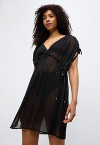 Μαύρο διάφανο φόρεμα κάλυψης με V-λαιμόκοψη, κοντά μανίκια, ρυθμιζόμενους δεσίματα στη μέση και διακοσμητικά στοιχεία σε χρυσούς τόνους. Ελαφρύ, αέρινο ύφασμα.