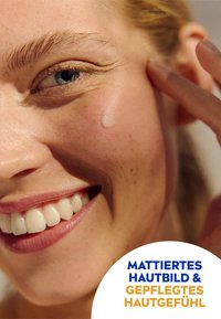 Nahaufnahme eines Frauenengesichts mit glatter Haut, einer subtilen glänzenden Textur und einem Wassertropfen auf ihrer Wange, der ein strahlendes Lächeln und klare Augen zeigt.