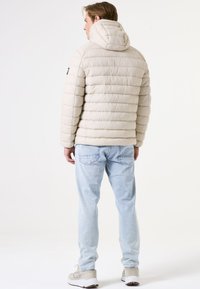 Beige Daunenjacke mit Kapuze, gestepptem Design, verstellbaren Zugbändern und seitlichen Taschen, kombiniert mit hellblauen Jeans und grauen Turnschuhen.