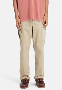 Beige cargo bukser med to store sidelommer, lige pasform og oprevne manchetter, parret med mørkebrune bådsko og en rød T-shirt.