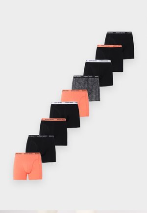 Αθλητικός νεαρός άνδρας στέκεται ξυπόλητος μπροστά σε λευκό τοίχο, φορώντας μαύρα και λευκά μποξεράκια με σχέδιο.